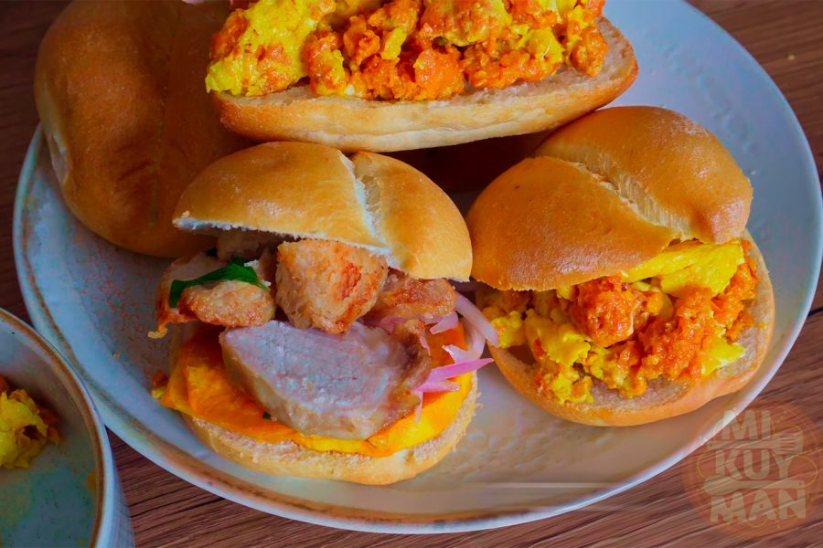 Pan con chicharrón peruano: crujientes chicharrones de cerdo sobre pan francés, camote frito y salsa criolla.