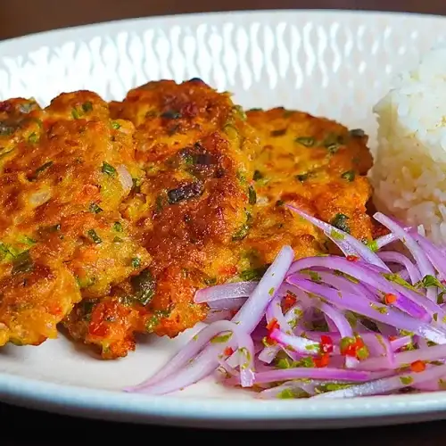 Torrejas de atún caseras, doradas y crujientes, servidas en un plato blanco con una guarnición de ensalada fresca de lechuga y tomate. Las tortitas muestran una textura crujiente por fuera y un interior suave y jugoso, con trozos visibles de atún y vegetales. La luz natural resalta los colores vibrantes de la comida