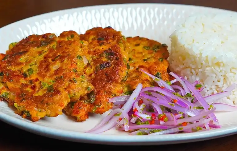 Torrejas de atún caseras, doradas y crujientes, servidas en un plato blanco con una guarnición de ensalada fresca de lechuga y tomate. Las tortitas muestran una textura crujiente por fuera y un interior suave y jugoso, con trozos visibles de atún y vegetales. La luz natural resalta los colores vibrantes de la comida