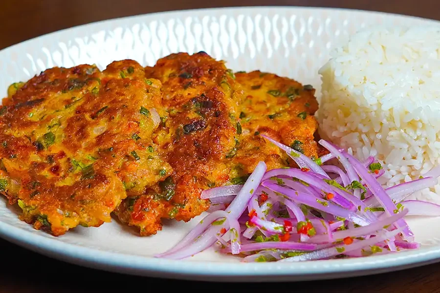 Torrejas de atún caseras, doradas y crujientes, servidas en un plato blanco con una guarnición de ensalada fresca de lechuga y tomate. Las tortitas muestran una textura crujiente por fuera y un interior suave y jugoso, con trozos visibles de atún y vegetales. La luz natural resalta los colores vibrantes de la comida